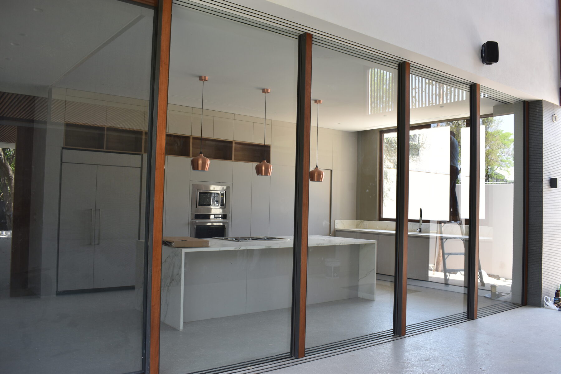 An open kitchen with a marble island and copper pendant lights, seen through timber-framed sliding glass.