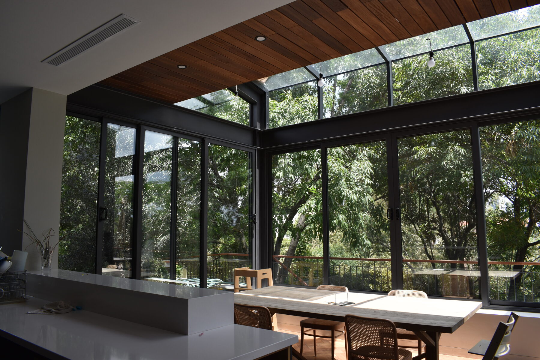 A glass dining pavilion with a timber ceiling, open on every side to the trees.