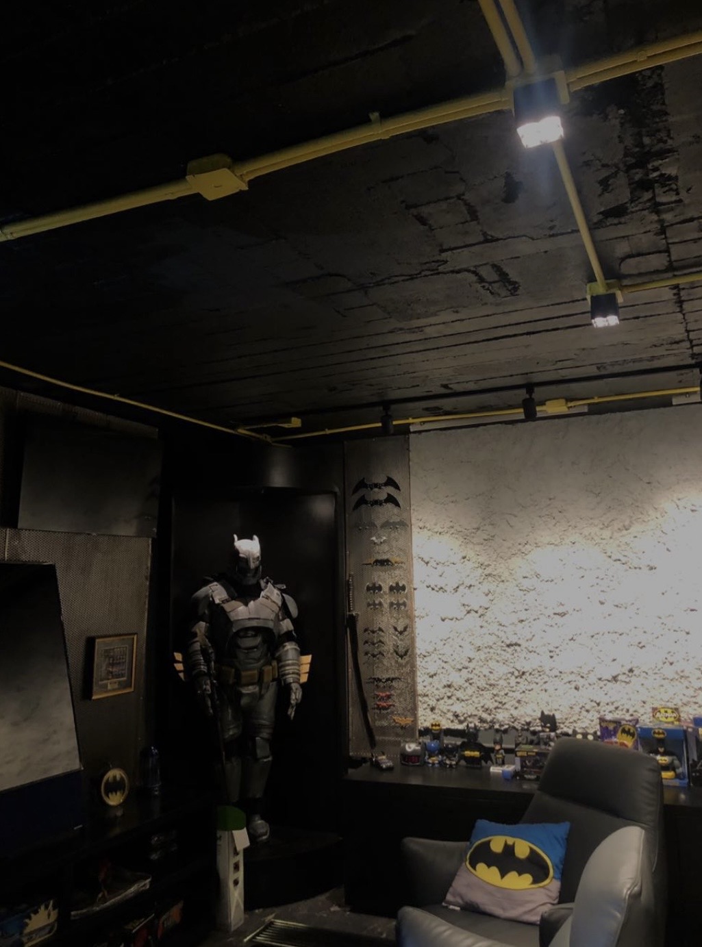 A dim collector's room with an armchair, a costumed figure, a textured stone wall and display shelves.